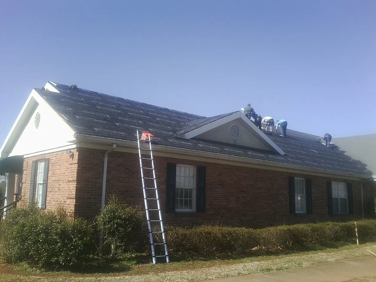 Crew tearing shingles off a brick ranch roof with a ladder set — Metal Roof Repair work by our crew in Erving